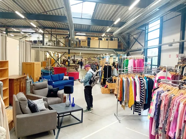 Ruim winkelbinnenaanzicht met kleding op rekken, meubels en bezoekers in kringloopwarenhuis