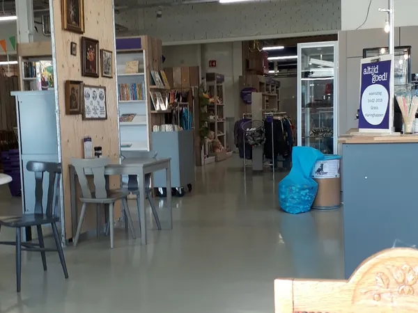 Winkelvloer met tafels, stoelen, stellingen en diversen spullen; blauwe afvalbak en balie zichtbaar