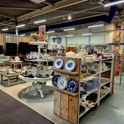 Ruim kringloopwinkelinterieur met huisraad, servies, meubilair en decoratieve items op verschillende stellingen en displays