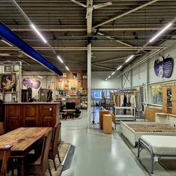 Ruim winkelinterieur met houten meubels, kleding op rekken, bedden en diverse tweedehands artikelen onder helderverlichting