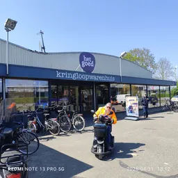Voorgevel van kringloopwinkel Het Goed met blauwe band, fietsen geparkeerd ervoor en klanten buiten