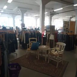 Winkelinterieur met kledingrekken vol tweedehands kleding, zithoek met stoelen en tafeltje in het midden