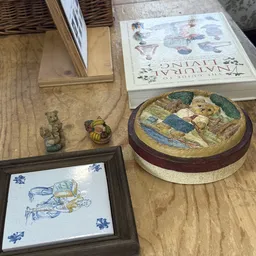 Uitgestalde tweedehands artikelen op houten tafel: kleine beeldjes, decoratief borduurwerk en een ronde decoratieve schaal me
