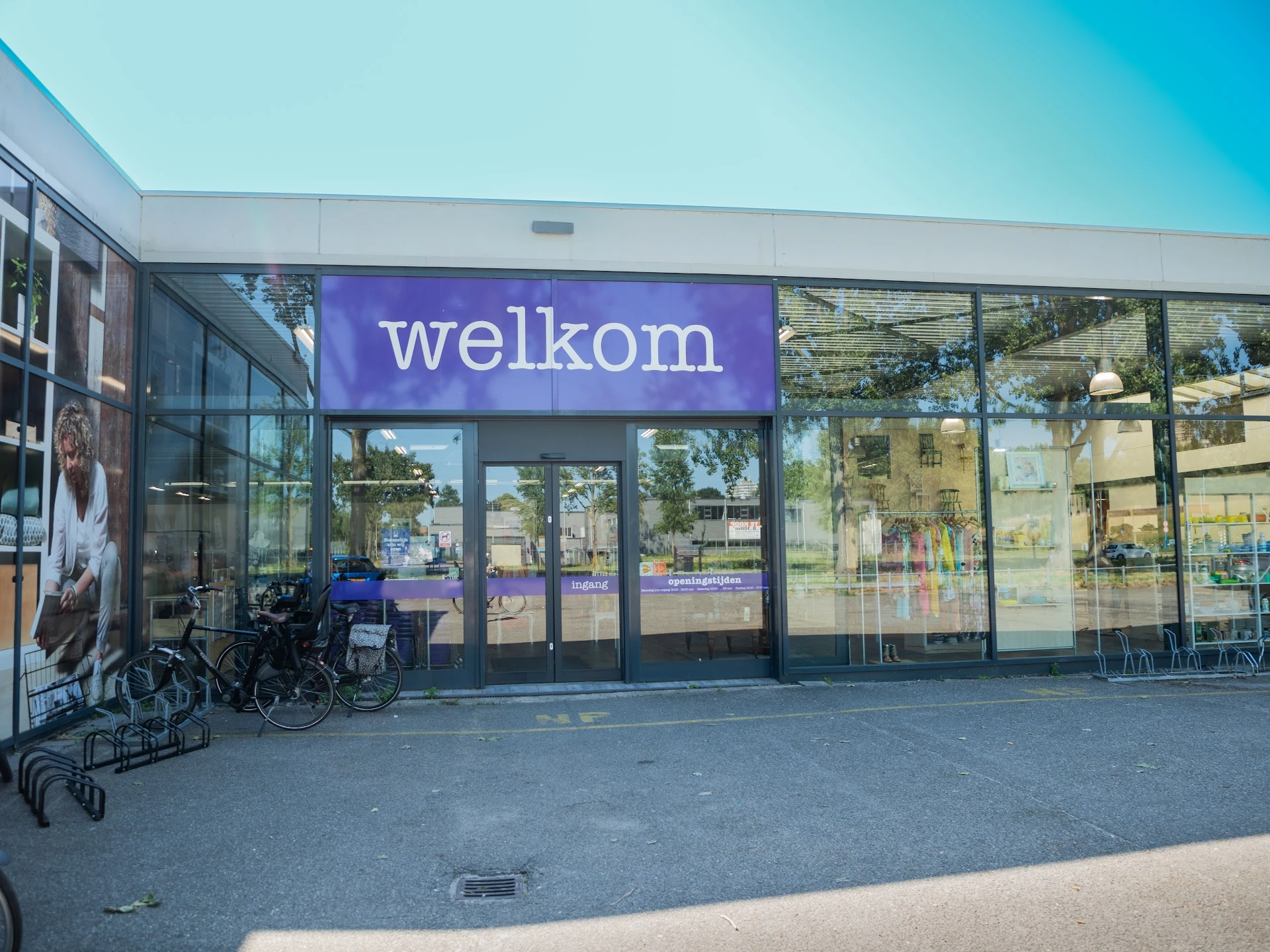 Gevel van kringloopwinkel met blauw 'welkom' logo, glazen deuren en ramen, winkeluitstalling zichtbaar