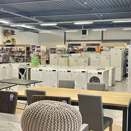 Uitgestalde wasmachines en drogers in een kringloopwinkel, met eettafel op de voorgrond