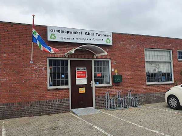 Bakstenen gevel van kringloopwinkel met bruine deur, twee ramen, uithangbord en Nederlandse vlag