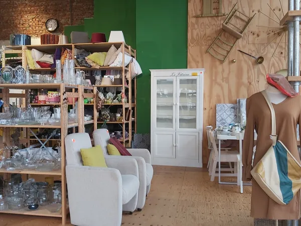 Kringloopwinkelinterieur met houten stellingen vol huishoudspullen, witte fauteuils, witte glazenkast, houten vloer en groene