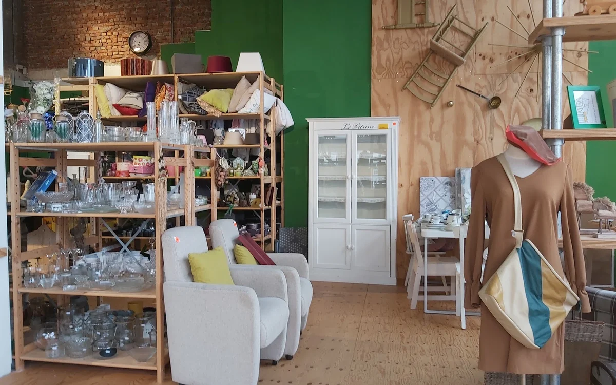 Kringloopwinkelinterieur met houten stellingen vol huishoudspullen, witte fauteuils, witte glazenkast, houten vloer en groene