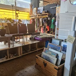 Winkelvloer met zitmeubels, kledingrekken, houten kasten en uitgestalde afbeeldingen in een kringloopwinkel