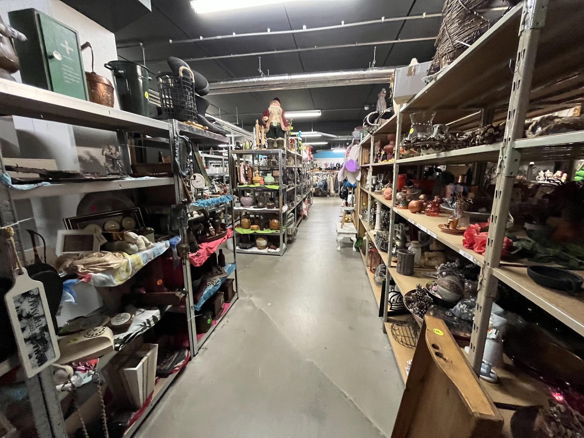 Winkelpad tussen metalen stellingen vol met tweedehands spullen, huishoudartikelen en diverse items in een kringloopwinkel.