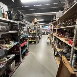 Winkelpad tussen metalen stellingen vol met tweedehands spullen, huishoudartikelen en diverse items in een kringloopwinkel.