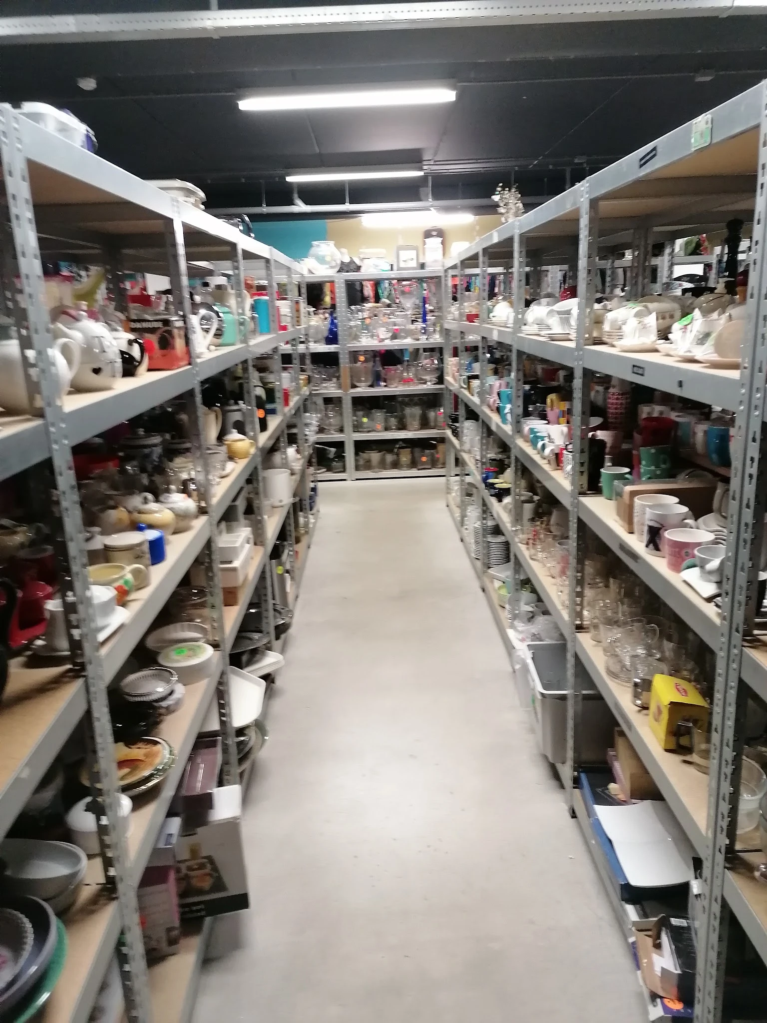 Lange gang tussen twee rijen metalen stellingen vol met huishoudelijke artikelen, serviesgoed en diversen in een kringloopwin