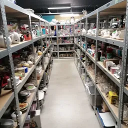 Lange gang tussen twee rijen metalen stellingen vol met huishoudelijke artikelen, serviesgoed en diversen in een kringloopwin