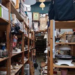 Nauwe gangpad tussen houten stellingen vol tweedehands spullen en kleding in kringloopwinkel
