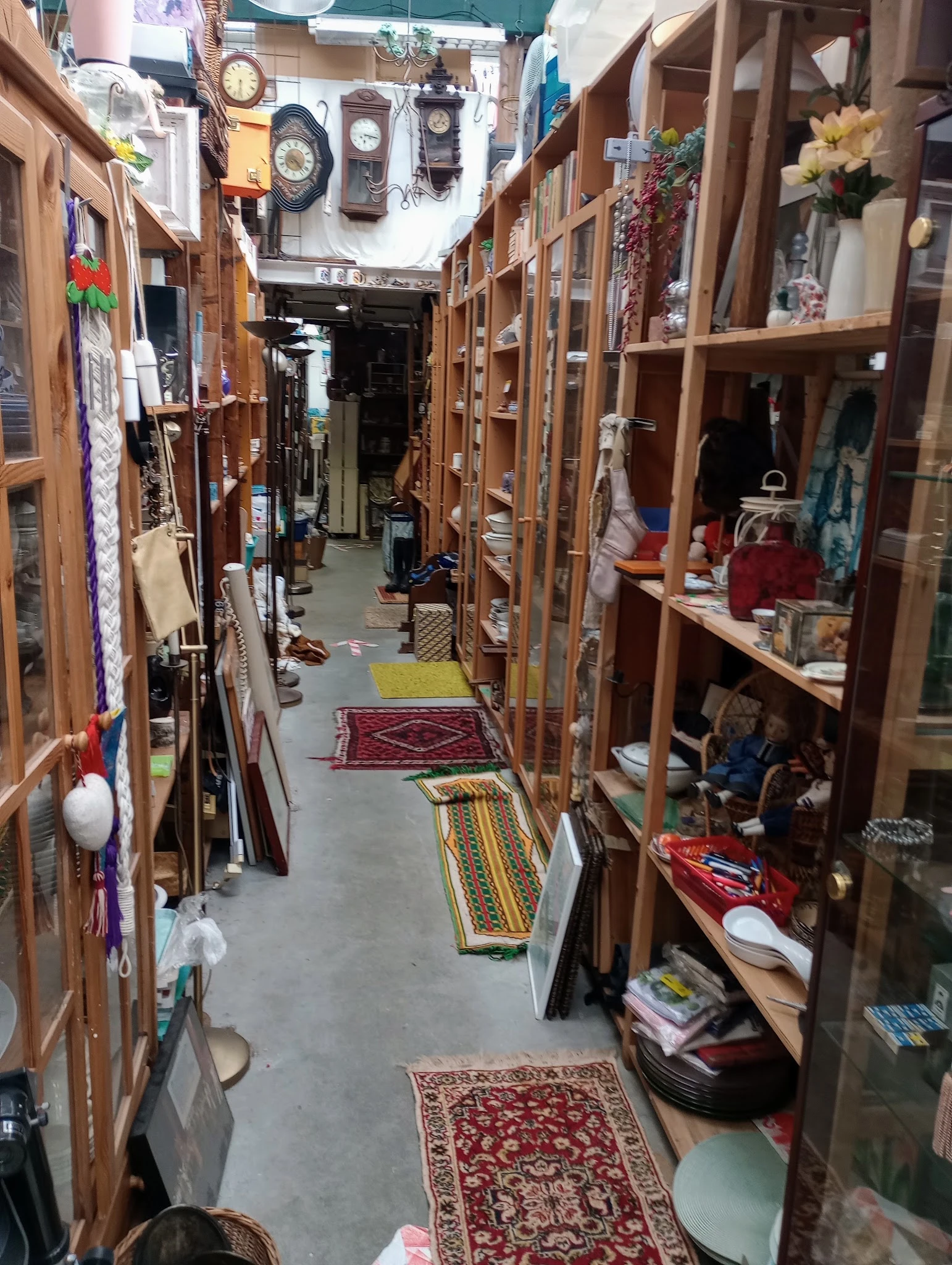 Lange gangpad in kringloopwinkel met houten kasten aan beide zijden, spullen op planken en kleine tapijten op de grond