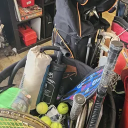 Verzameling sportartikelen waaronder tennisracketten, golfballen en diverse tassen in een kringloopwinkel