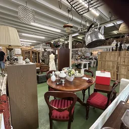 Interieur van kringloopwinkel met houten tafel, rode stoelen, kasten, lampen en diverse meubels en huishoudartikelen.