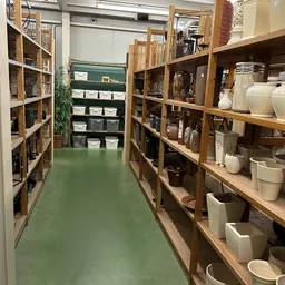 Smalle winkelgang met houten stellingen vol huishoudelijke spullen en witte serviesgoed aan beide zijden, groen linoleum vloe