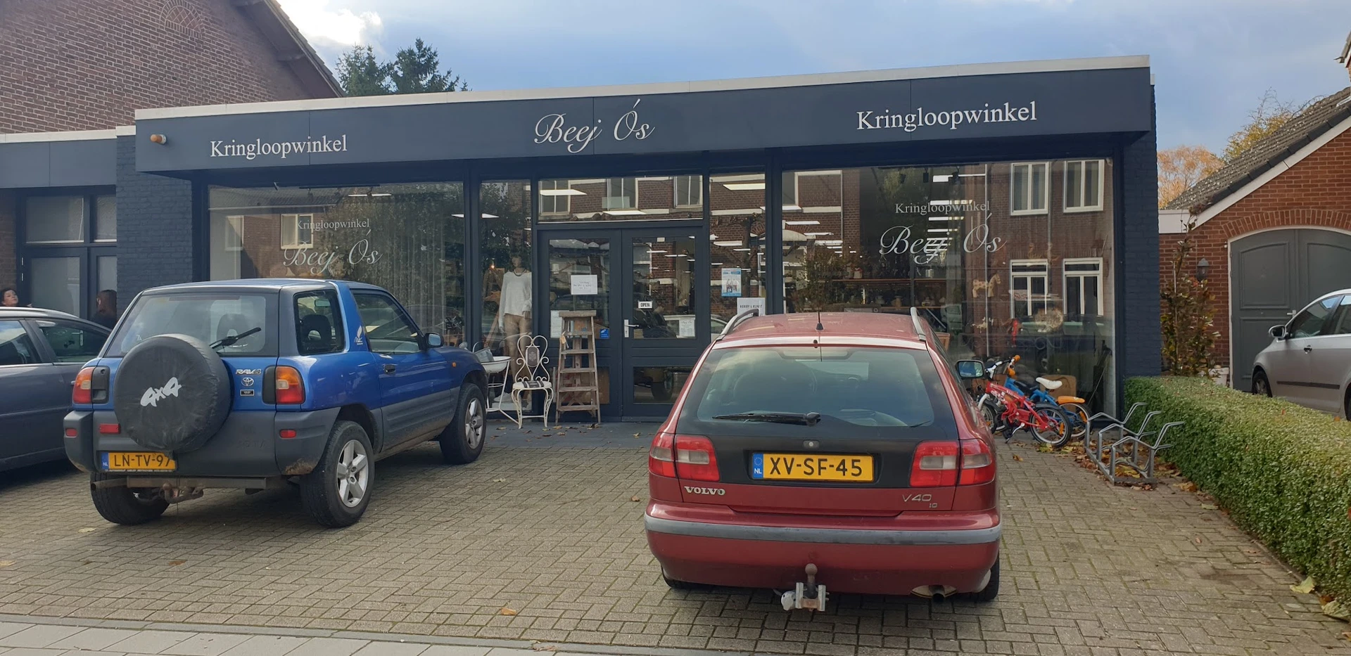 Voorgevel van kringloopwinkel Beej Ós met donkerblauwe pui, winkelnaam zichtbaar, parkeerplaats met auto's ervoor