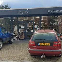 Voorgevel van kringloopwinkel Beej Ós met donkerblauwe pui, winkelnaam zichtbaar, parkeerplaats met auto's ervoor