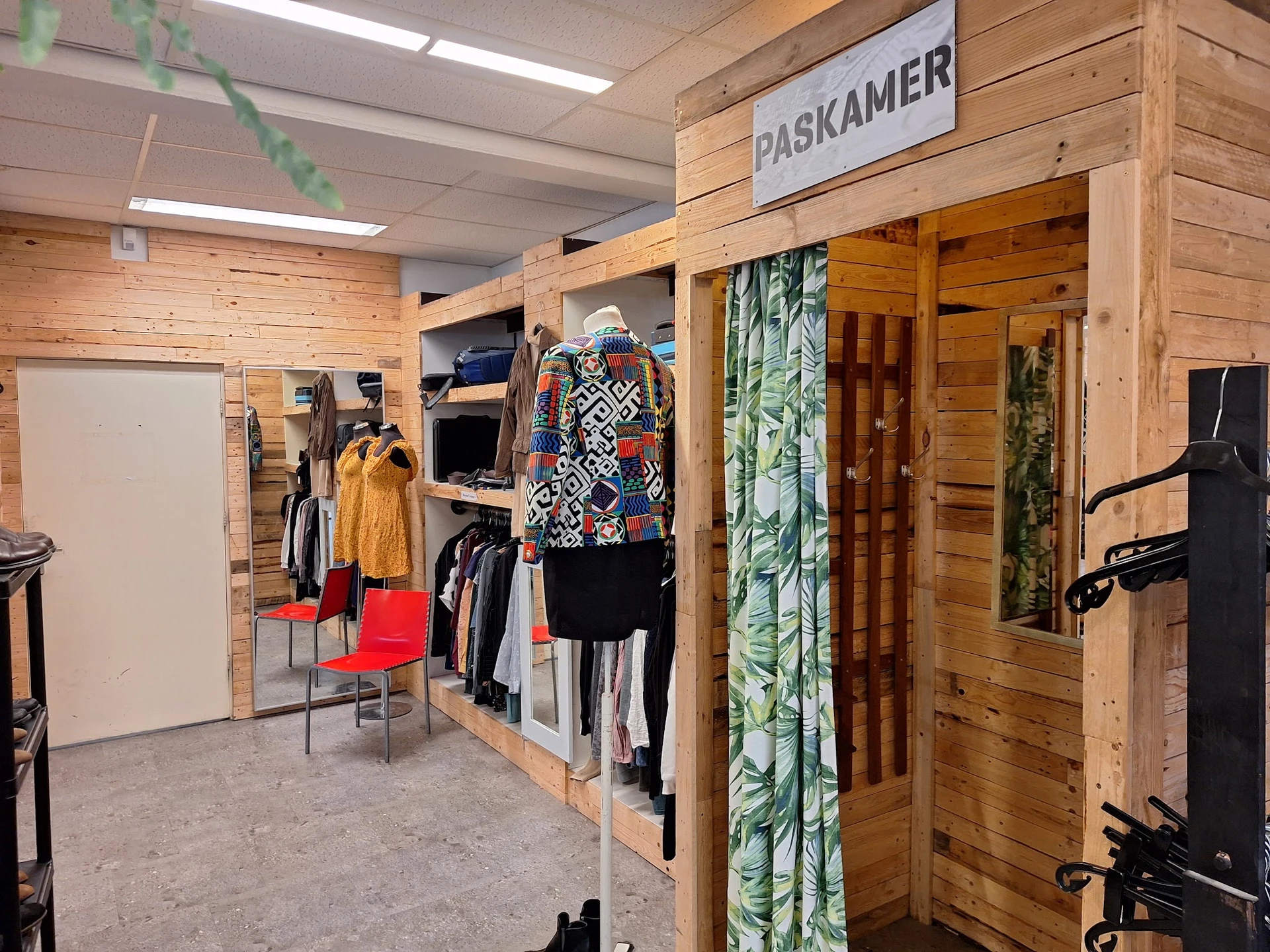 Winkelvloer met houten boxen, kledingrekken en uitgestalde tweedehands kleding, waaronder een gele jas en kleurrijke kledings