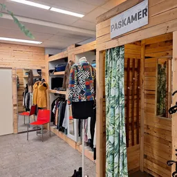 Winkelvloer met houten boxen, kledingrekken en uitgestalde tweedehands kleding, waaronder een gele jas en kleurrijke kledings