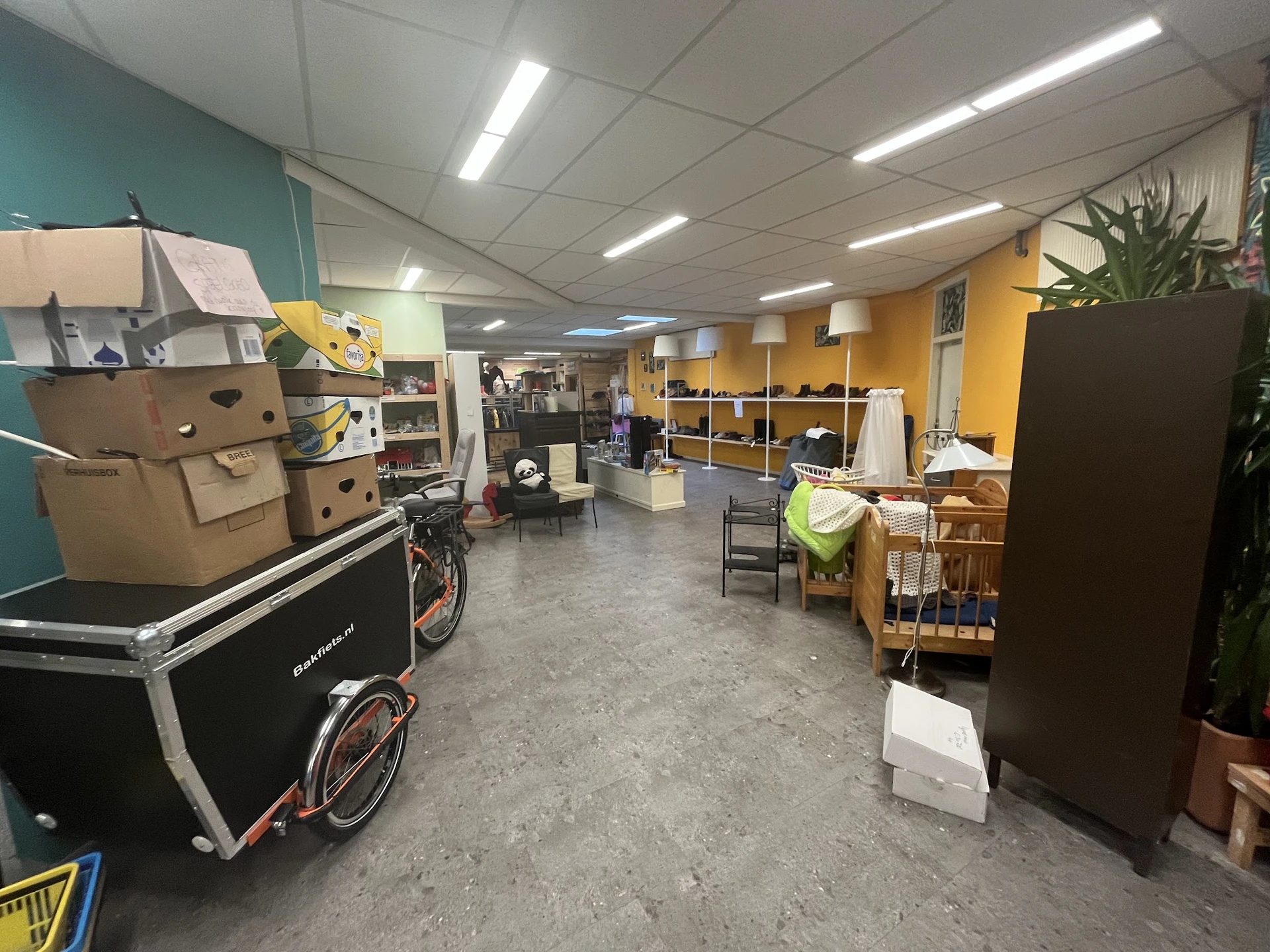 Winkelvloer van kringloopwinkel met stellingen, dozen, fietsen en diverse spullen tegen gekleurde wanden