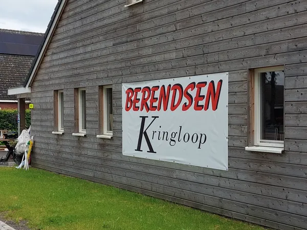 Grijze houten gevel van kringloopwinkel met wit uithangbord met rode en zwarte letters, groene voortuin