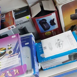 Diverse elektronica en technische apparaten in dozen op een tafel, waaronder scanners en andere randapparatuur.