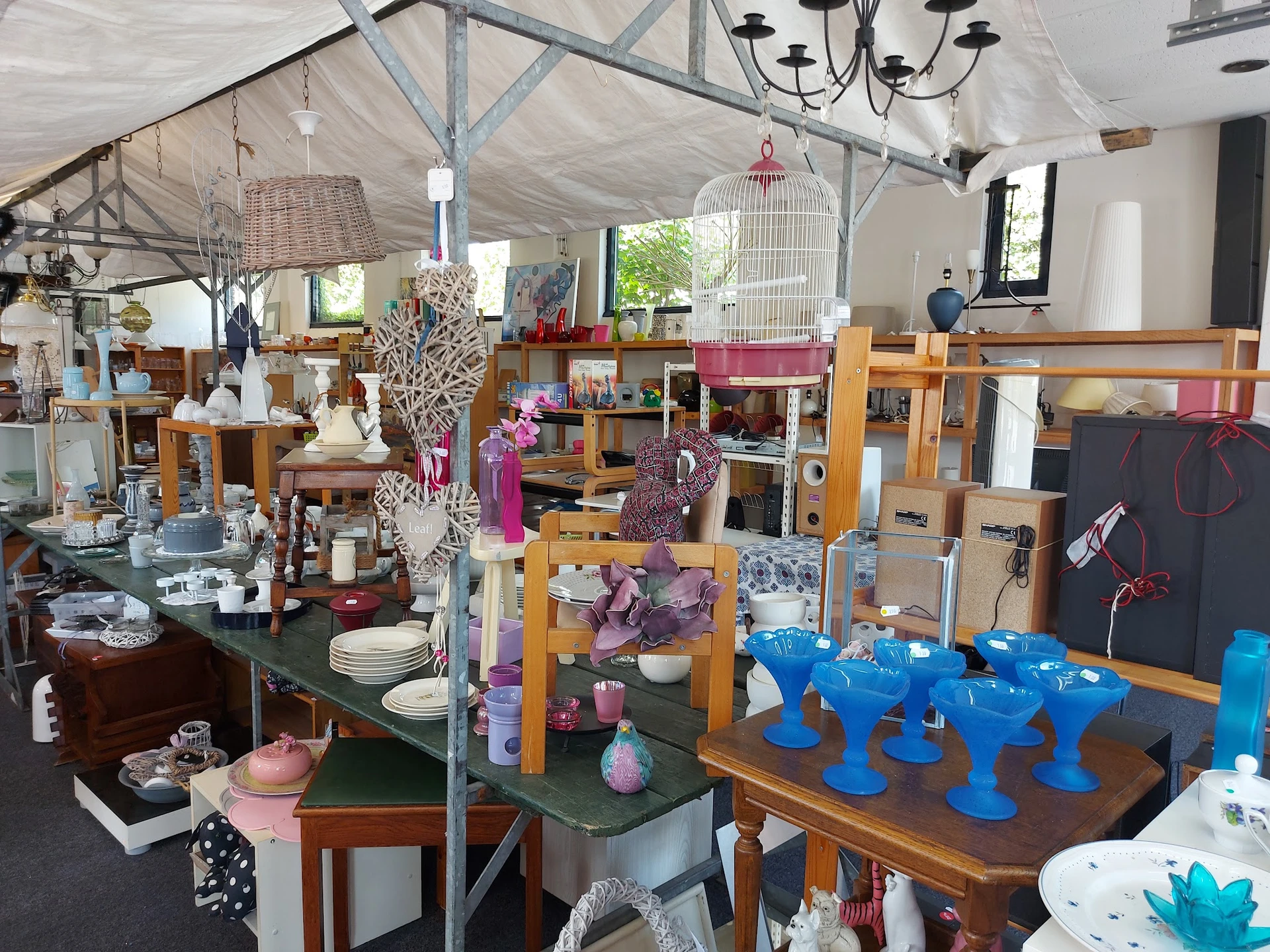 Winkelbinnenruimte met veel uitgestalde spullen op tafels en stellingen, waaronder servies, decoraties en meubelen.