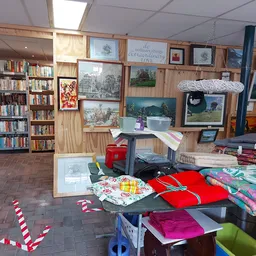 Winkelbinnenruimte met boekenkasten, kunstwerken aan muur, textielwaren en huishoudartikelen op tafels