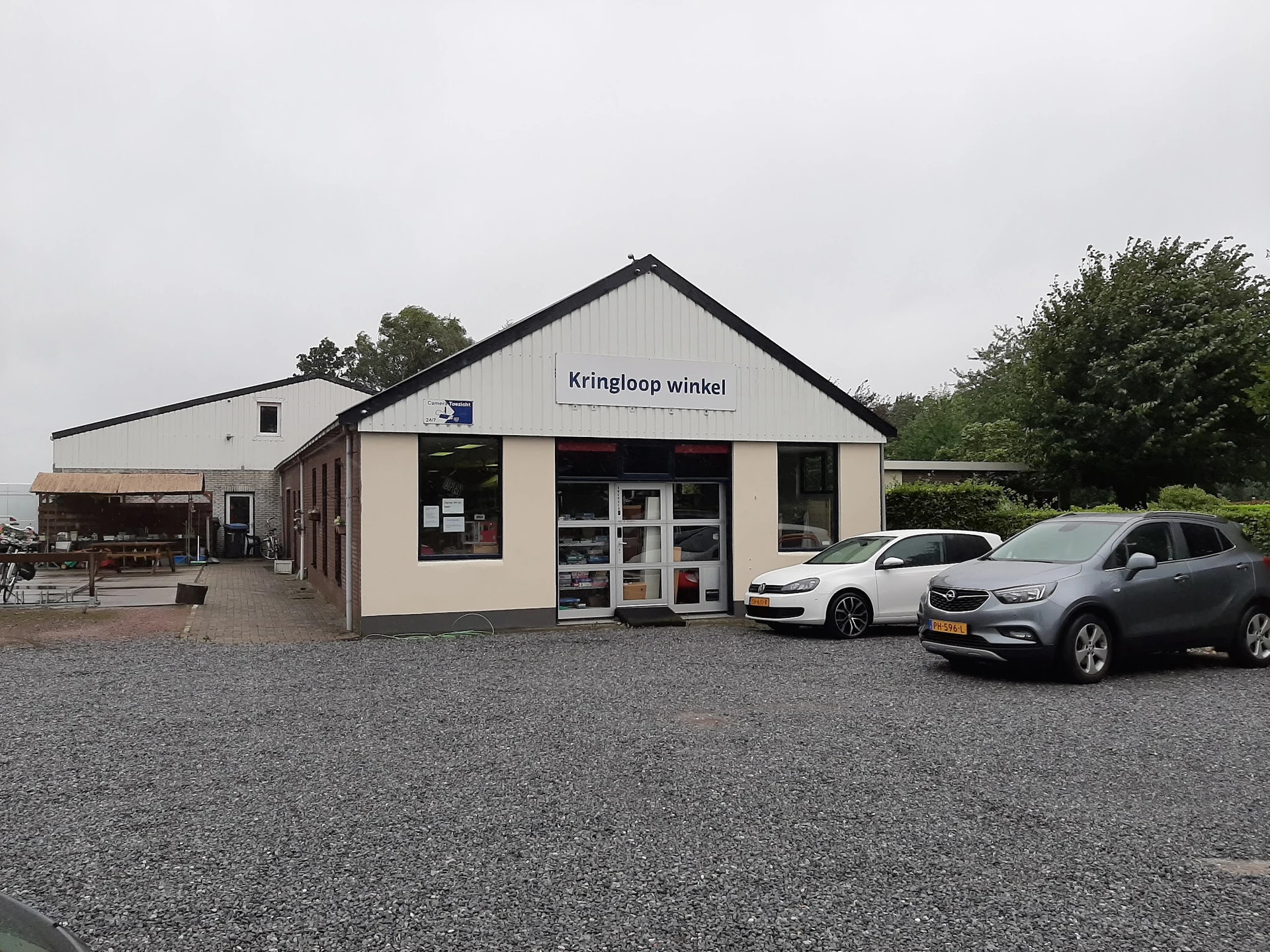 Witte gevel van kringloopwinkel met zwak dak, naam 'Kringloop winkel' op voorgevel, twee auto's geparkeerd op grind