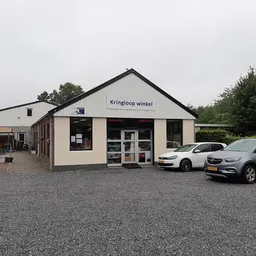 Witte gevel van kringloopwinkel met zwak dak, naam 'Kringloop winkel' op voorgevel, twee auto's geparkeerd op grind