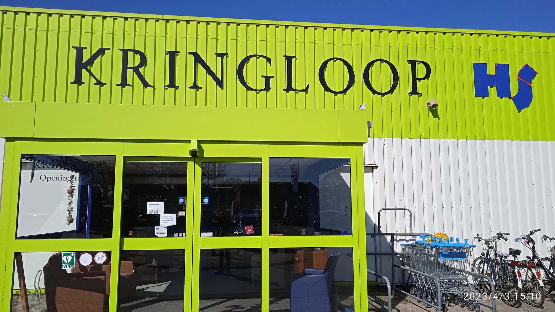 Geel-groene gevel van kringloopwinkel met grote ramen en winkeldeuren, winkelwagentjes buiten