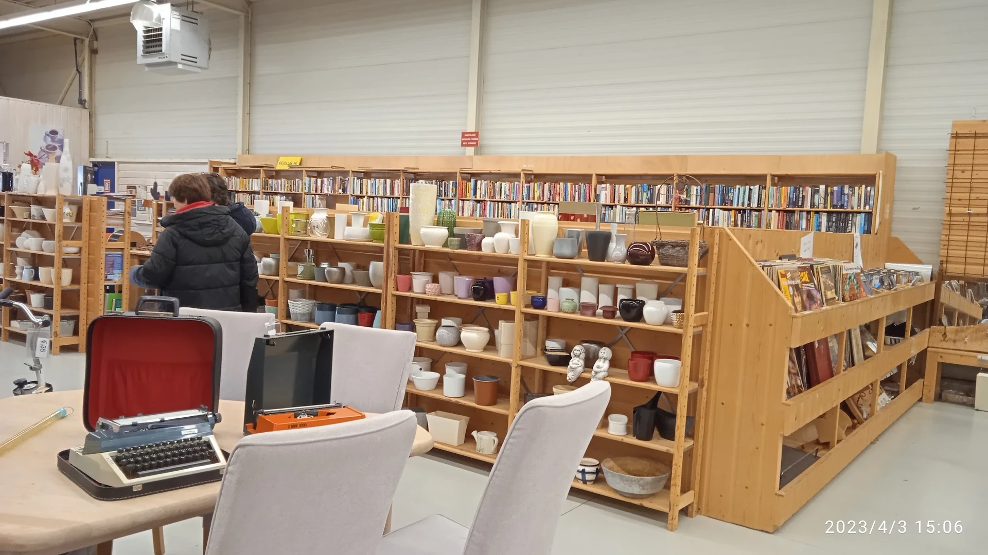 Binnenaanzicht van kringloopwinkel met houten stellingen vol boeken, serviesgoed en huishoudartikelen, laptop op tafel op voo