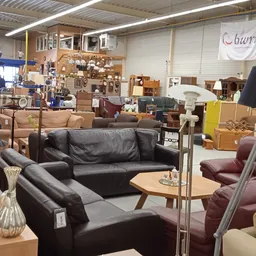 Winkelvloer met uitgestalde meubels: zwarte lederen bank, rode leunstoel en houten tafel, met stellingen en dozen op de achte