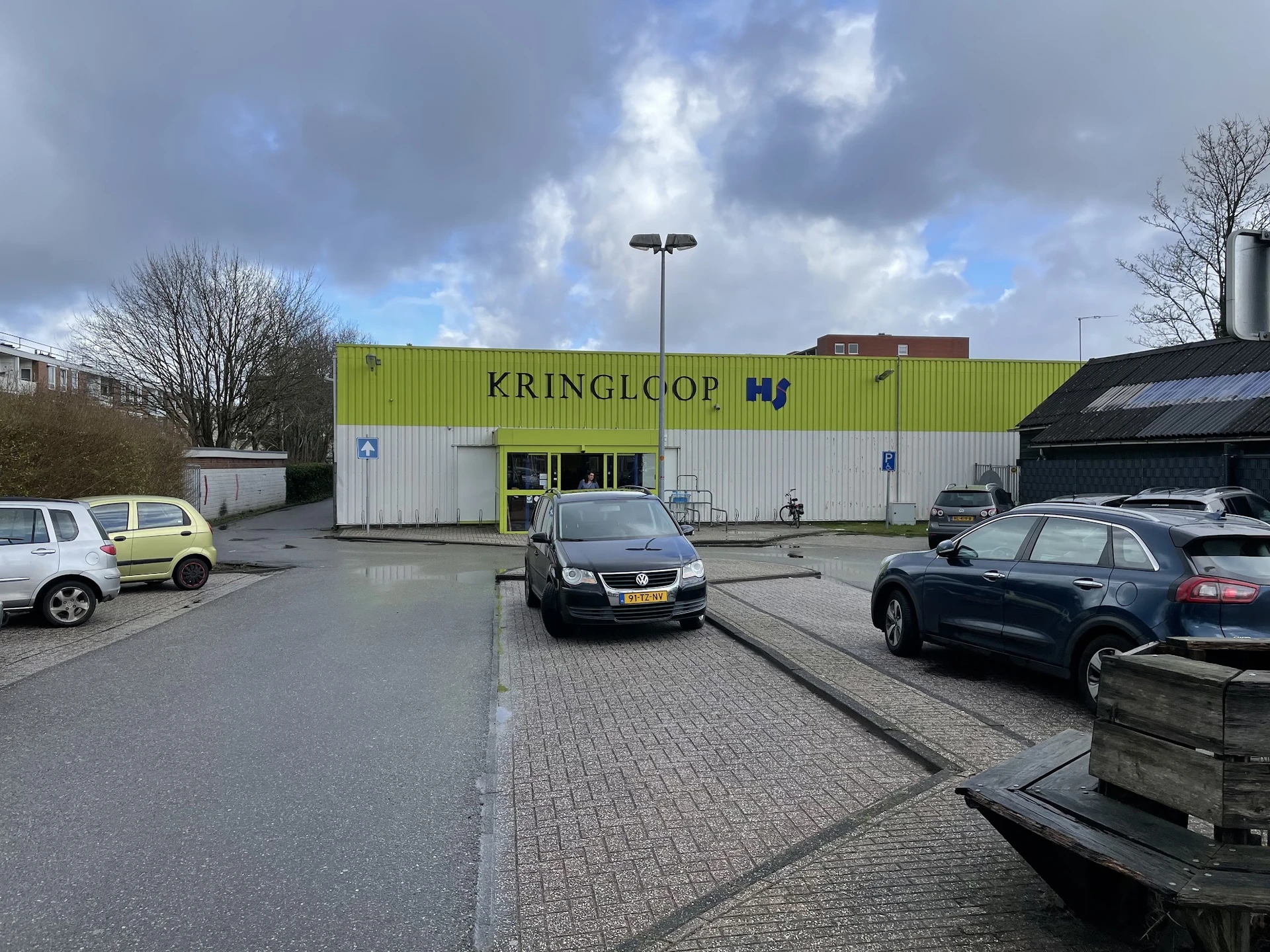 Geel-groen golfkartonnen gevel van kringloopwinkel Kringlcop met winkelraam en deur, geparkeerde auto's en straatverlichting