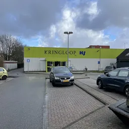 Geel-groen golfkartonnen gevel van kringloopwinkel Kringlcop met winkelraam en deur, geparkeerde auto's en straatverlichting