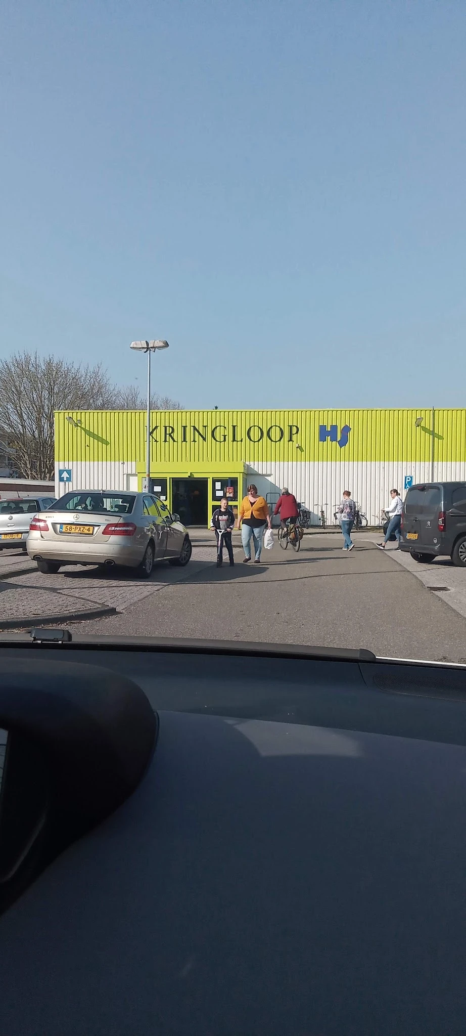 Gele gevel van kringloopwinkel Kringloop met parkeerterrein en bezoekers voor de ingang