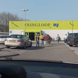 Gele gevel van kringloopwinkel Kringloop met parkeerterrein en bezoekers voor de ingang