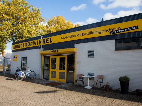 Witte gevel van kringloopwinkel met geel accent, zwarte naam 'Kringloopwinkel' en gele deuren, winkelnaam en logo duidelijk z