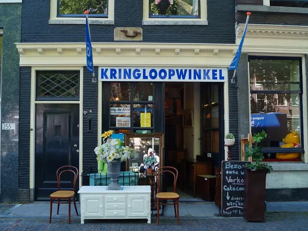 Gevel van kringloopwinkel met blauwe luifel, naam KRINGLOOPWINKEL zichtbaar, twee stoelen en bloemen voor de ingang