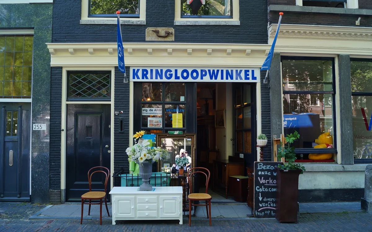 Voorgevel van kringloopwinkel met blauwe luifel, winkelnaam op het raam, open deur en bloemen voor de winkel