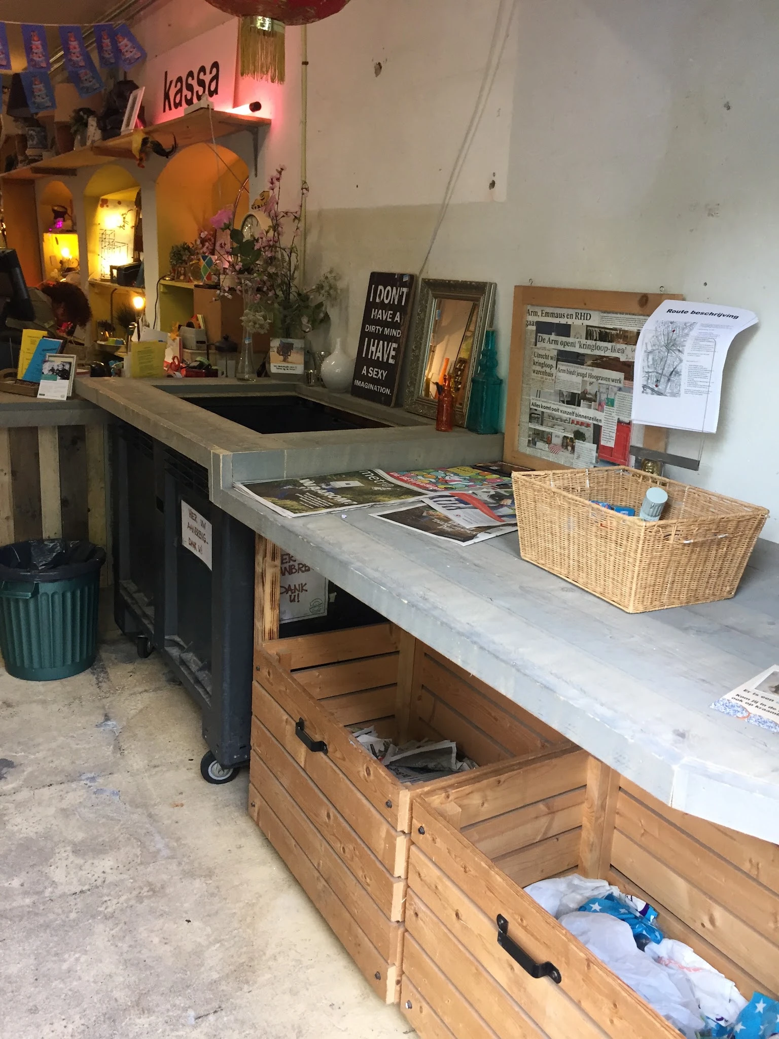 Werkplek in kringloopwinkel met toonbank, opbergmanden, kranten en diverse spullen op en onder het werkblad