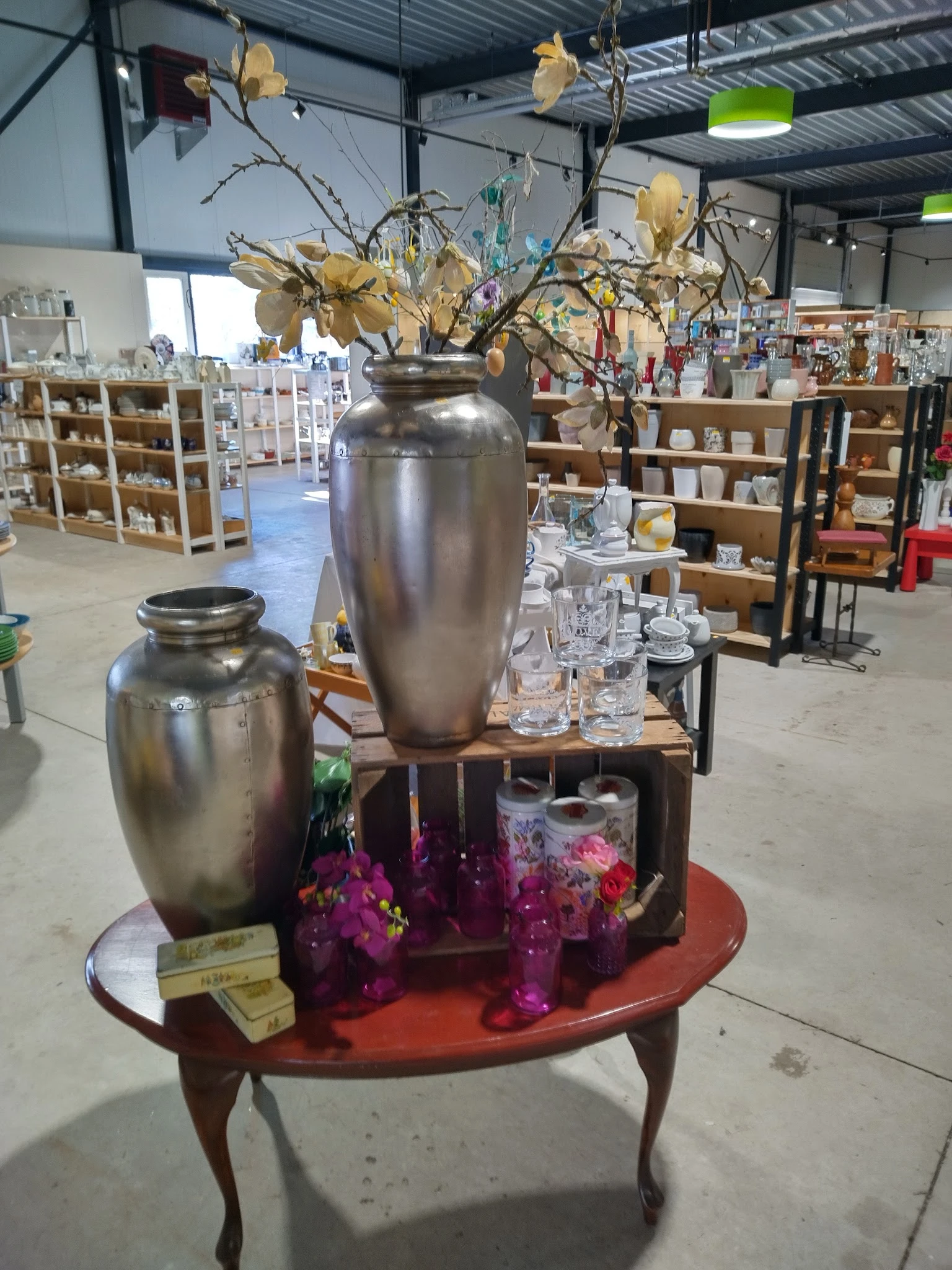 Twee zilveren urnen met bloemstukken en glazen op een houten tafel in de kringloopwinkel