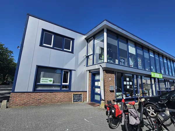 Moderne gevel van kringloopwinkel met witte bovenbouw en bakstenen onderbouw, grote ramen, blauwe deur en fietsen geparkeerd 