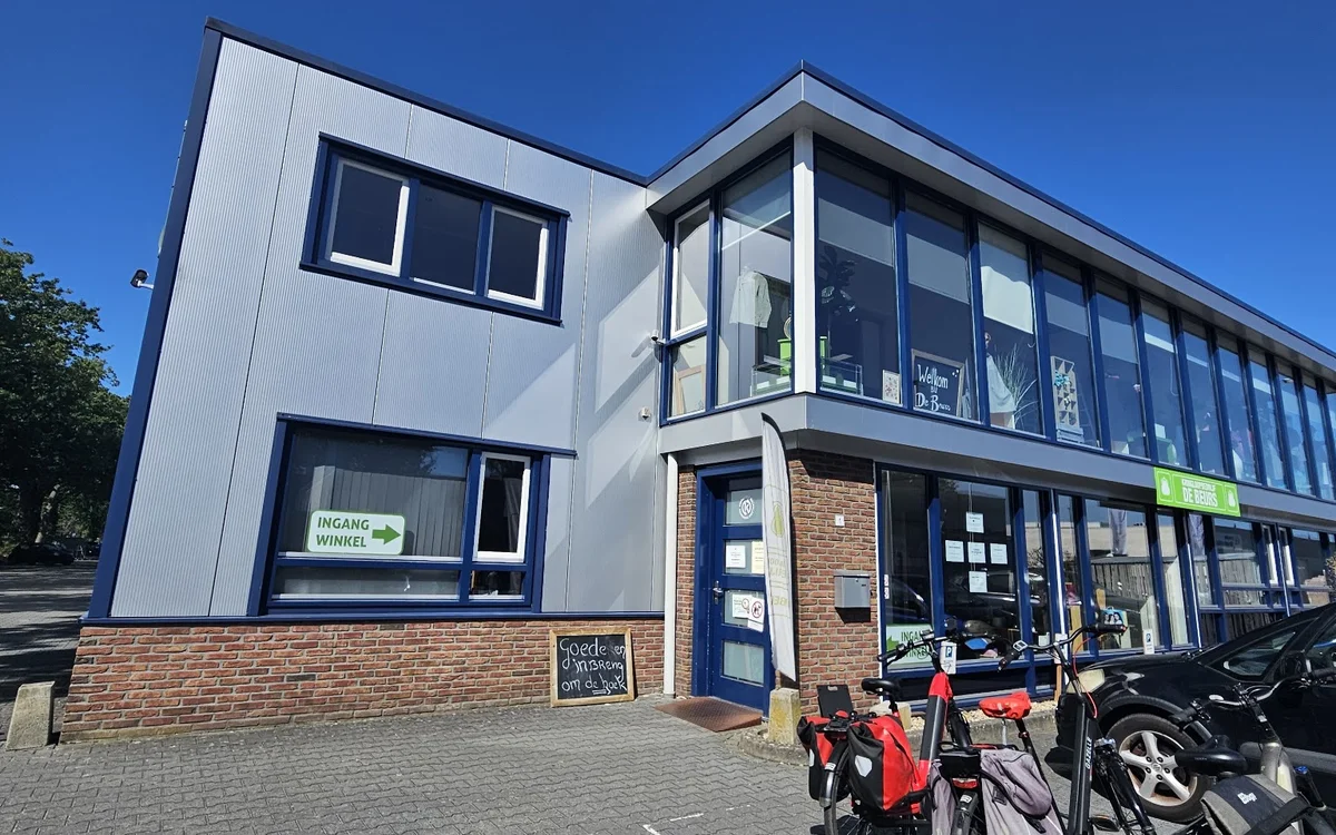 Moderne gevel van kringloopwinkel met witte bovenbouw en bakstenen onderbouw, grote ramen, blauwe deur en fietsen geparkeerd 