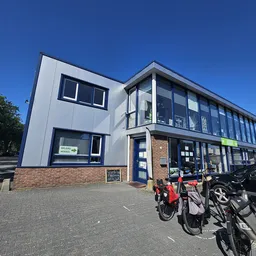 Moderne gevel van kringloopwinkel met witte bovenbouw en bakstenen onderbouw, grote ramen, blauwe deur en fietsen geparkeerd 