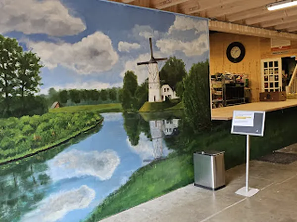 Groot muurschildering van Nederlandse windmolen aan water in kringloopwinkel, met houten kassa-area op achtergrond