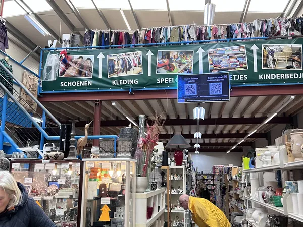 Overdekt winkelinterieur met kleding aan rekken en aan het plafond, en diverse spullen op tafels en stellingen.